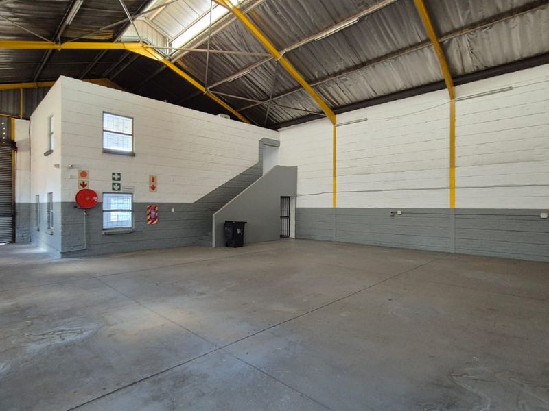 Spacious industrial warehouse unit featuring high ceilings with exposed steel trusses, concrete flooring, a roller shutter door, and an internal staircase.