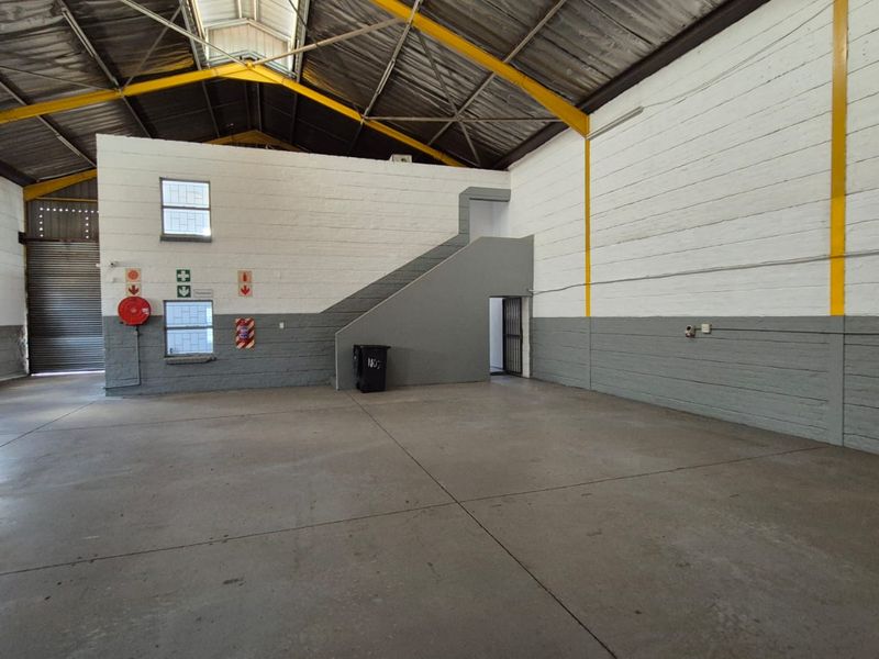 Spacious industrial warehouse unit featuring high ceilings with exposed steel trusses, concrete flooring, a roller shutter door, and an internal staircase.