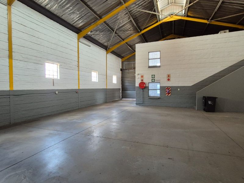 Spacious industrial warehouse unit featuring high ceilings with exposed steel trusses, concrete flooring, and a large roller shutter door for easy access.