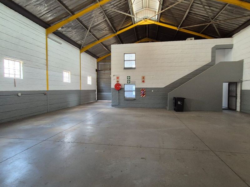 Spacious industrial warehouse unit featuring high ceilings with exposed steel trusses, concrete flooring, a roller shutter door, and an internal staircase.