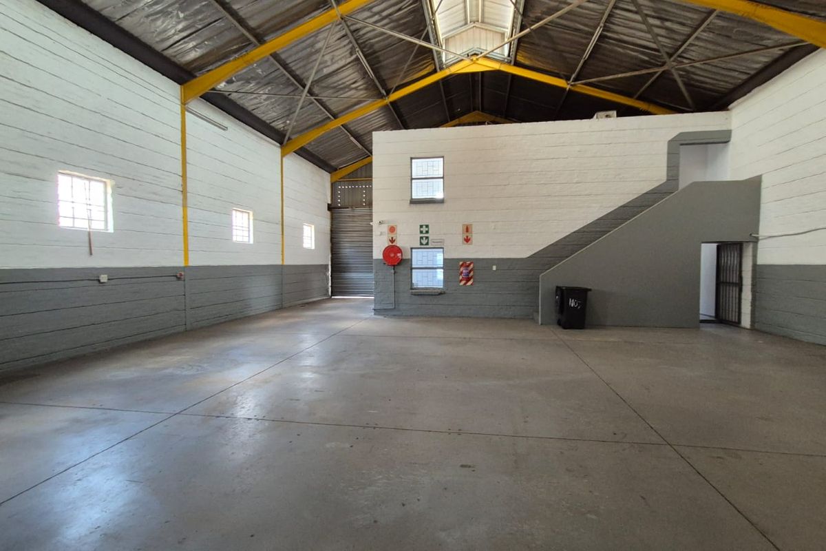 Spacious industrial warehouse unit featuring high ceilings with exposed steel trusses, concrete flooring, a roller shutter door, and an internal staircase.