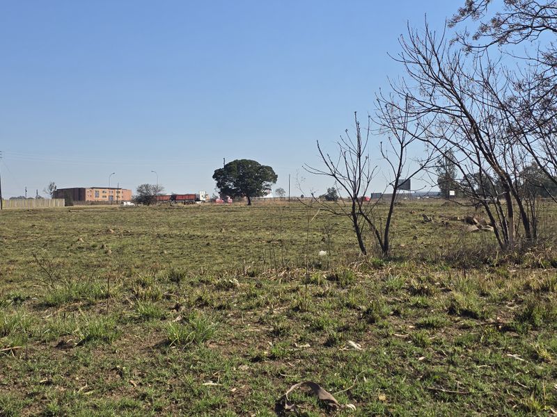 A wide-angle view of a vacant commercial land plot featuring natural grass cover and mature trees, suitable for future development.