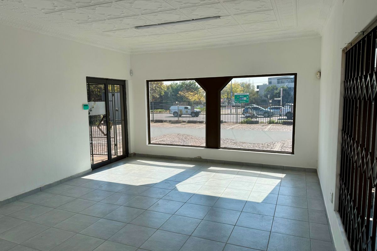 A vacant commercial office space featuring tiled flooring, decorative ceiling tiles, and a large street-facing display window with security gates.