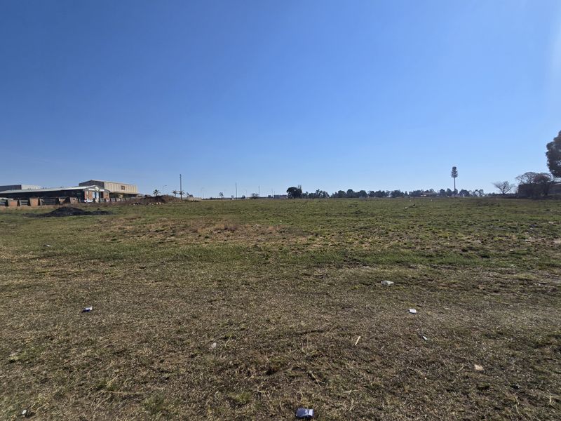 A wide-angle view of a large, flat, vacant commercial plot of land with grass cover and surrounding industrial buildings in the distance.