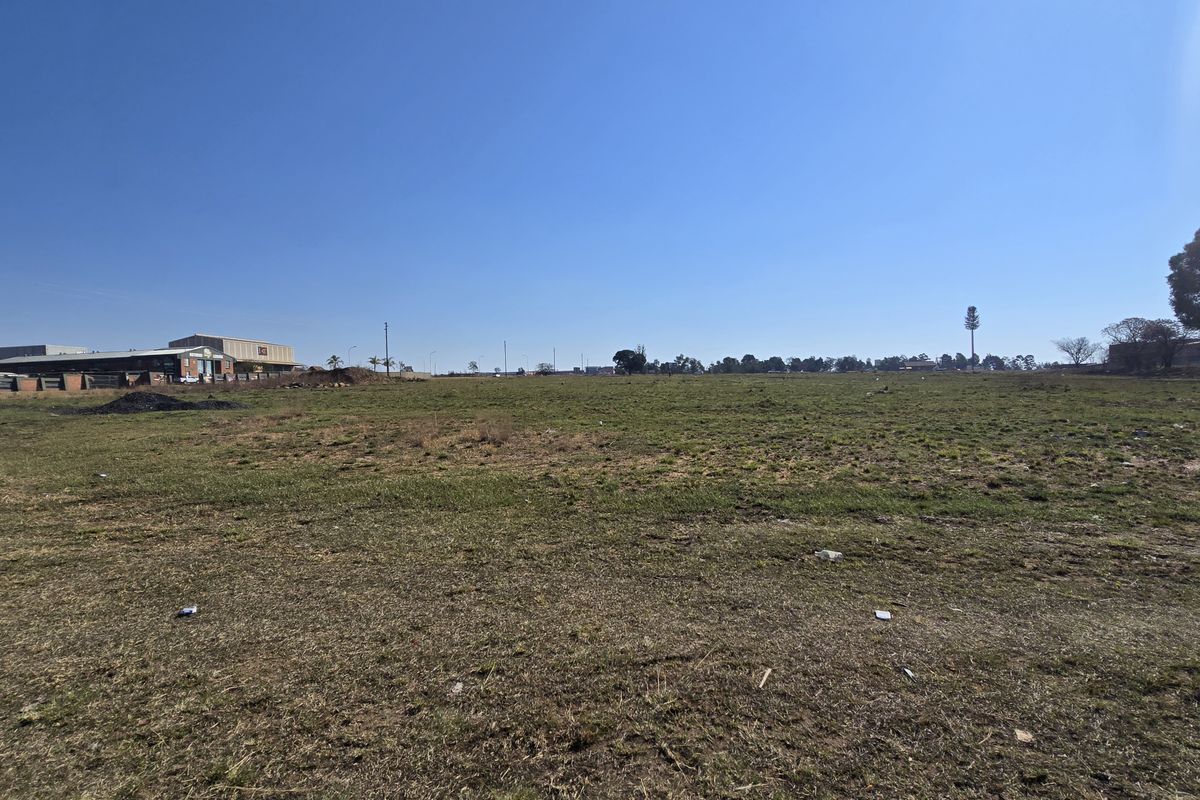 A wide-angle view of a large, flat, vacant commercial plot of land with grass cover and surrounding industrial buildings in the distance.