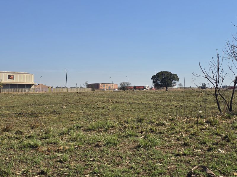 A wide-angle view of a vacant commercial land plot featuring open grass fields, perimeter fencing, and surrounding industrial infrastructure in the distance.