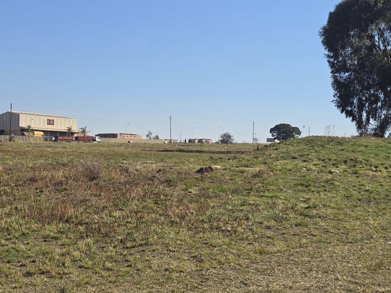 A wide-angle view of an undeveloped commercial land plot featuring natural grassland and surrounding industrial infrastructure in the distance.