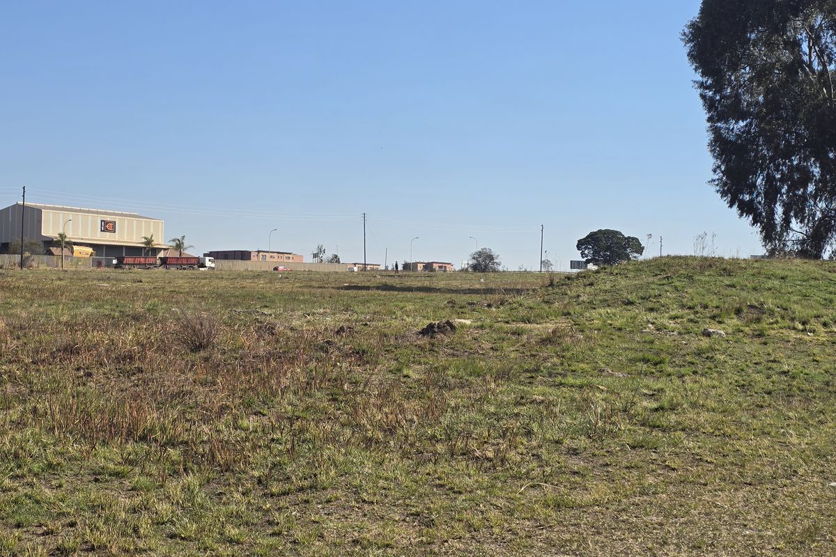 A wide-angle view of an undeveloped commercial land plot featuring natural grassland and surrounding industrial infrastructure in the distance.