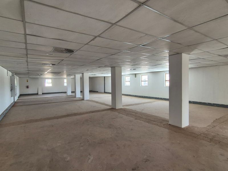 A large, open-plan office space featuring a suspended ceiling, structural columns, and multiple windows, currently in a vacant, shell-like condition.