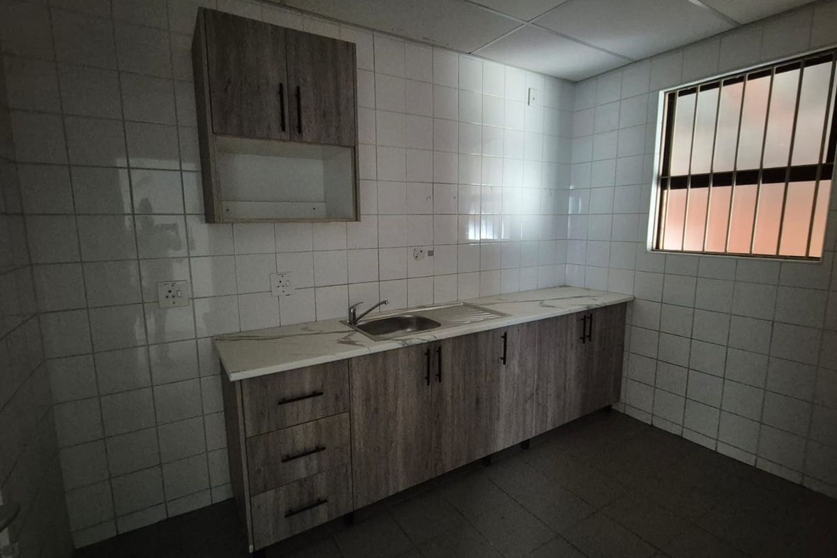 A functional office kitchenette featuring wood-look cabinetry, a stainless steel sink, tiled walls, and a window with security bars.