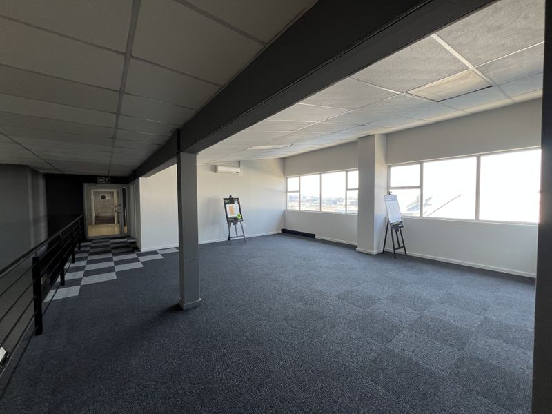 A spacious, open-plan office area featuring carpeted flooring, a suspended ceiling, and large windows providing ample natural light and views.