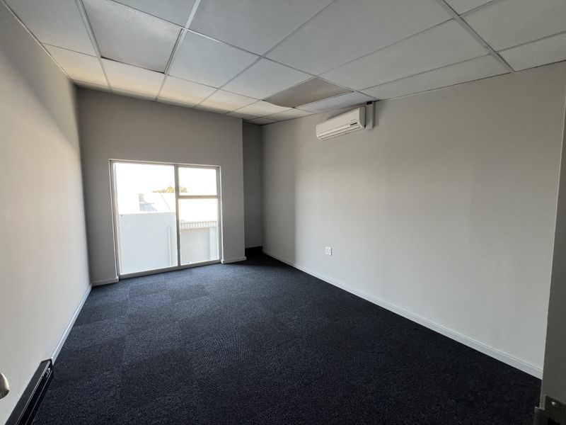 A clean, empty office space featuring dark carpet tiles, a suspended ceiling, and a wall-mounted split-system air conditioning unit.