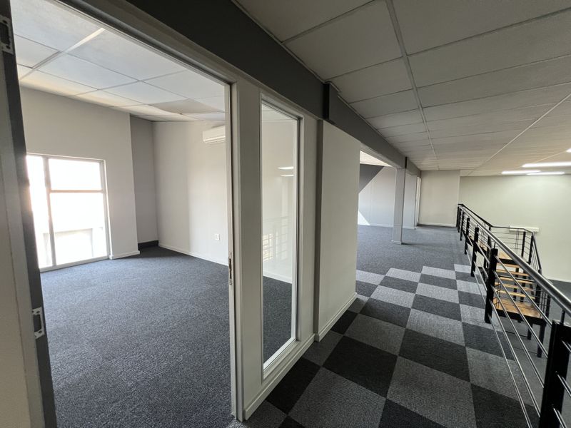 Modern office space featuring a private office room, open-plan mezzanine area, carpeted flooring, and glass partitioning with a staircase.