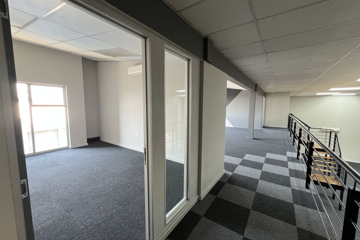 Modern office space featuring a private office room, open-plan mezzanine area, carpeted flooring, and glass partitioning with a staircase.
