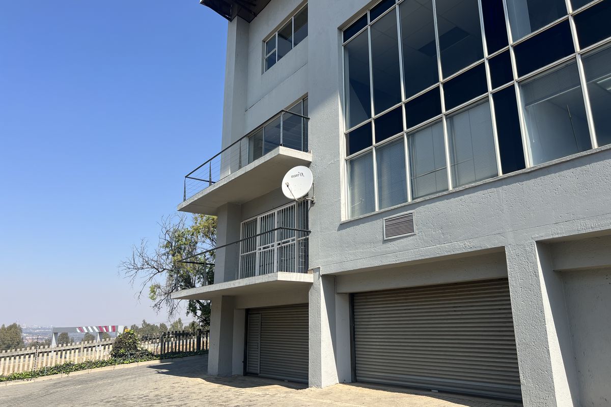 Rear elevation of a multi-story office building featuring roller shutter garage doors, balconies with glass railings, and large glass facade panels.