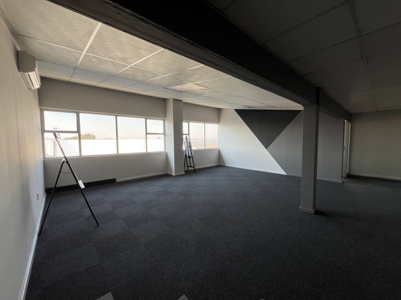 Spacious open-plan office area featuring large windows, carpeted flooring, a suspended ceiling, and a wall-mounted air conditioning unit.