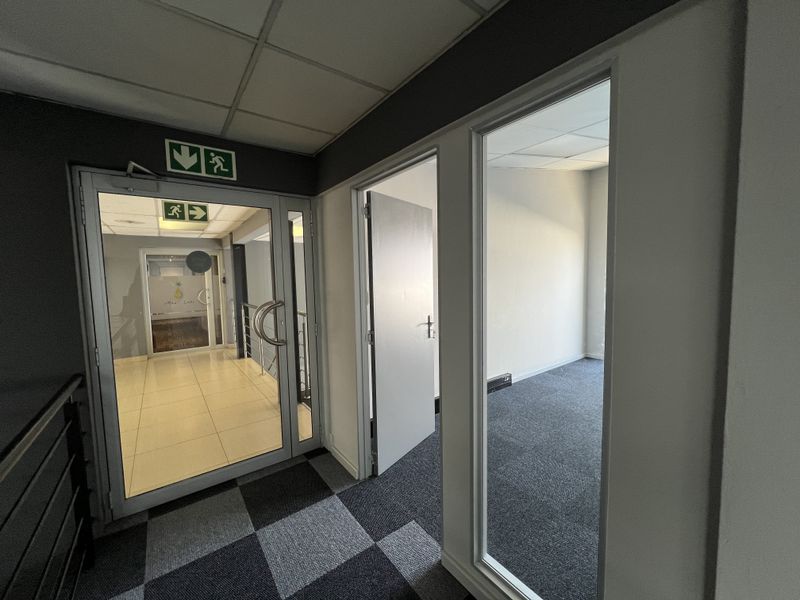 A clean office hallway featuring carpeted flooring, suspended ceilings, and glass-partitioned office entrances with clear emergency exit signage.
