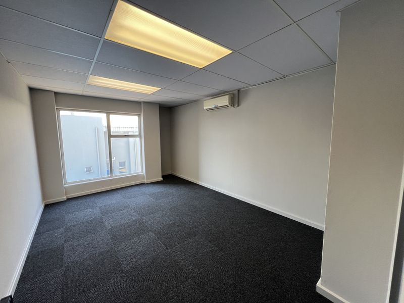 A clean, empty office space featuring dark carpet tiles, a suspended ceiling with recessed lighting, and a wall-mounted air conditioning unit.