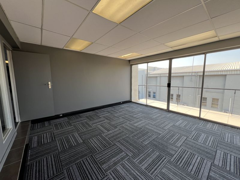 A bright, empty office space featuring modern carpet tiles, a suspended ceiling with recessed lighting, and large windows leading to a balcony.