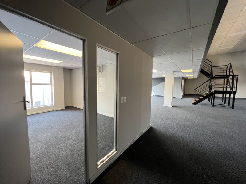 Spacious open-plan office area featuring carpeted flooring, a modern metal staircase, suspended ceilings with recessed lighting, and a private glass-partitioned office space.