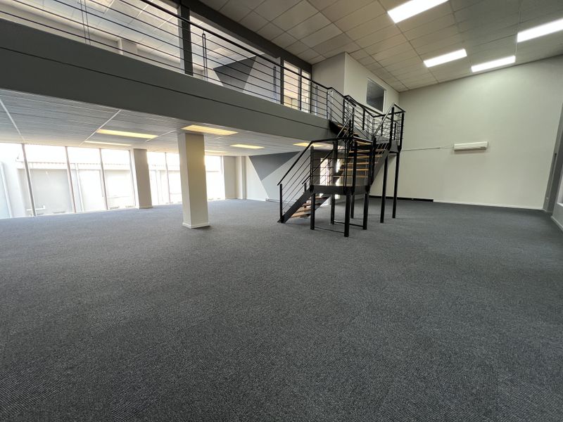 Spacious open-plan office area featuring a mezzanine level, modern metal staircase, carpeted flooring, and large windows providing ample natural light.