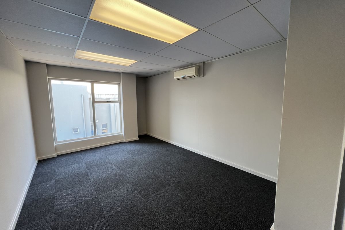 A clean, empty office space featuring dark carpet tiles, a suspended ceiling with recessed lighting, and a wall-mounted air conditioning unit.
