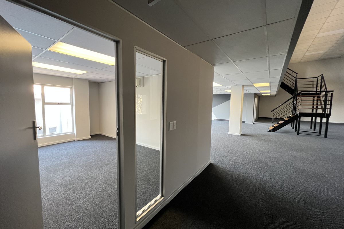 Spacious open-plan office area featuring carpeted flooring, a modern metal staircase, suspended ceilings with recessed lighting, and a private glass-partitioned office space.