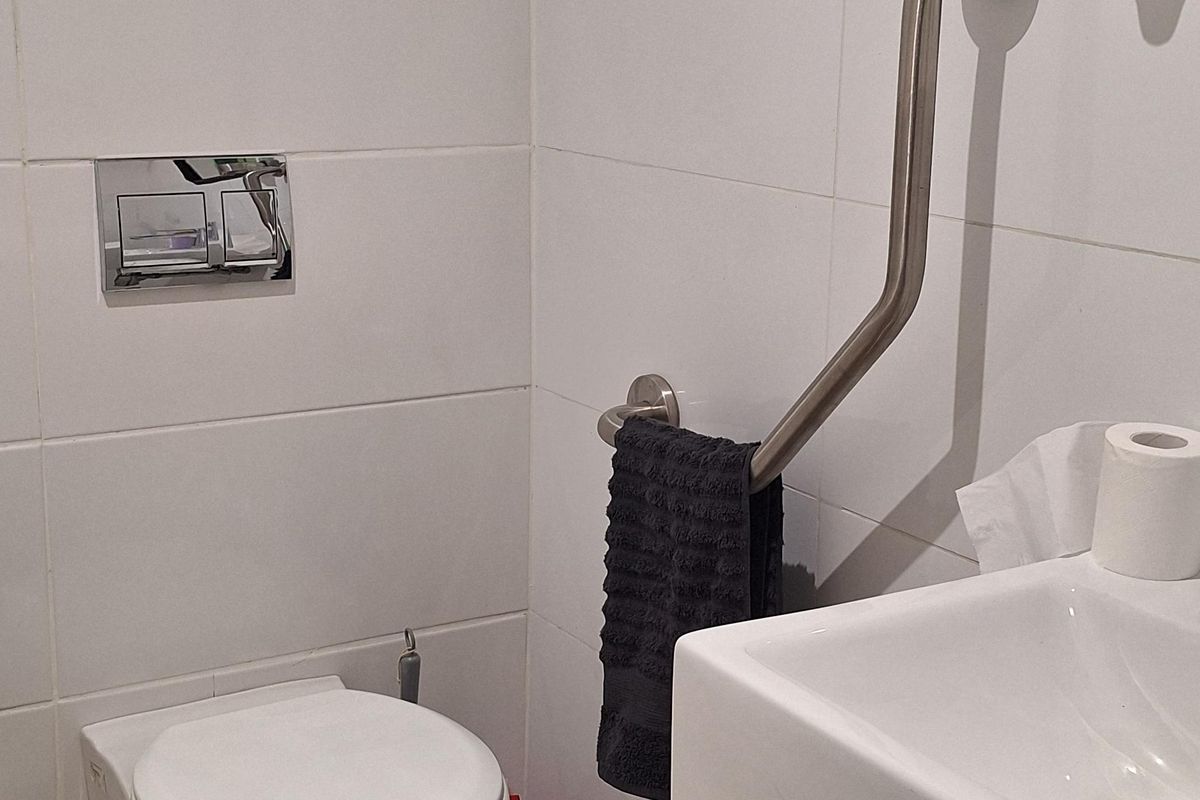 A functional commercial bathroom featuring a wall-mounted toilet, wall-mounted sink, and an accessible grab rail with white tiled walls.