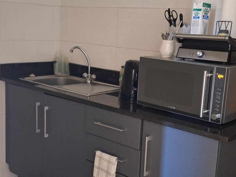 A compact kitchenette featuring grey cabinetry, a stainless steel sink, granite countertops, and a tiled backsplash with exposed plumbing.