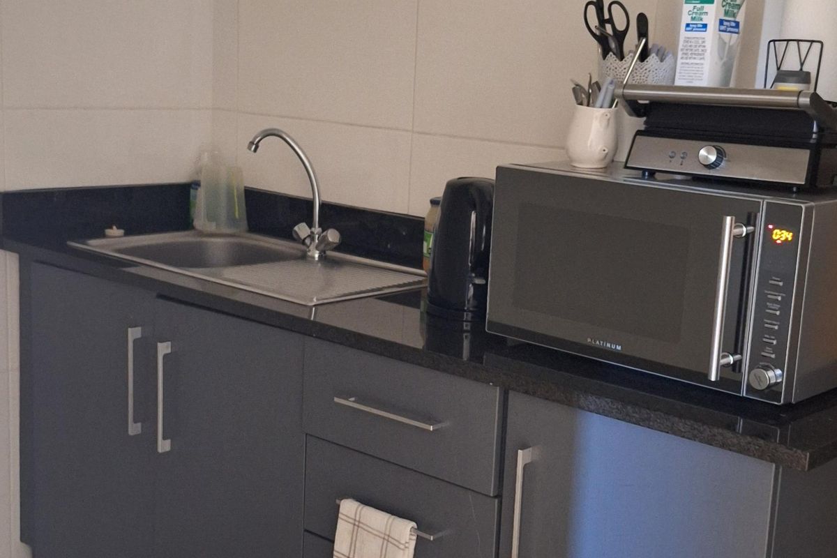 A compact kitchenette featuring grey cabinetry, a stainless steel sink, granite countertops, and a tiled backsplash with exposed plumbing.