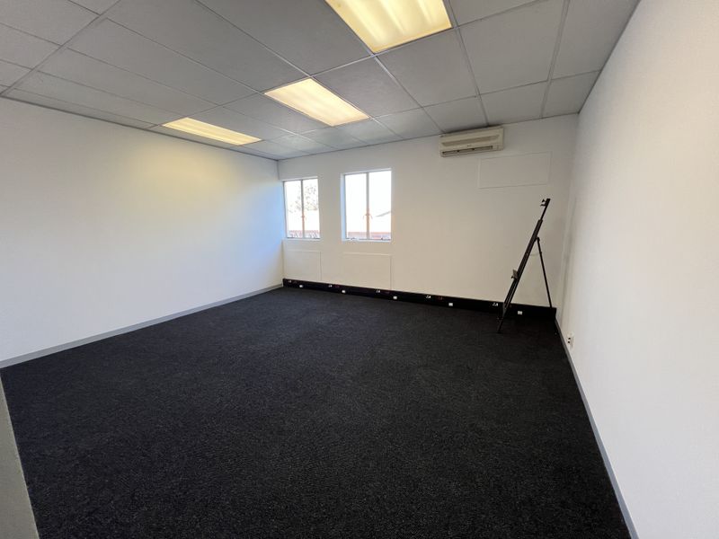 A clean, vacant office space featuring dark carpeting, a suspended ceiling with recessed lighting, and a wall-mounted air conditioning unit.