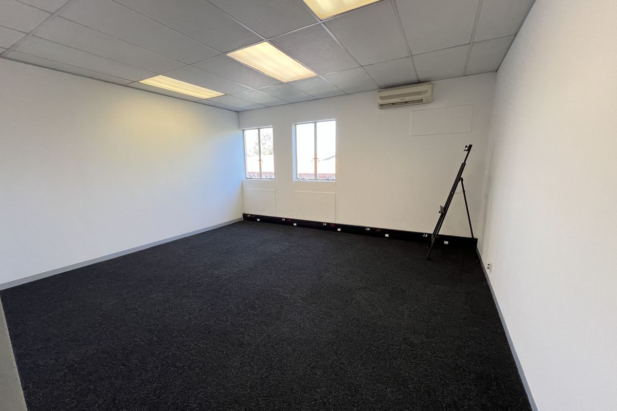 A clean, vacant office space featuring dark carpeting, a suspended ceiling with recessed lighting, and a wall-mounted air conditioning unit.