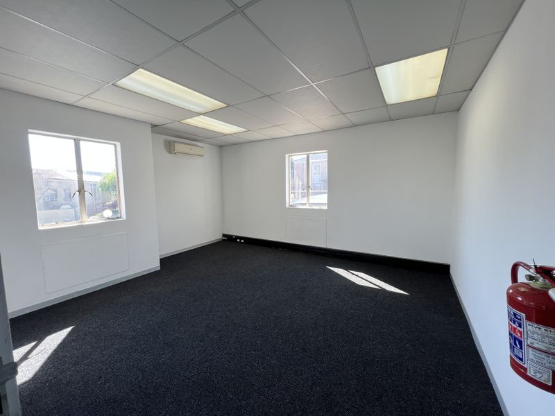 A clean, vacant office space featuring dark carpeting, a suspended ceiling with recessed lighting, and a wall-mounted split-system air conditioner.