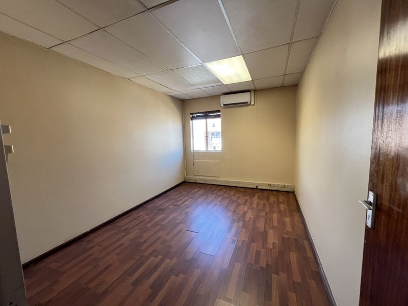A vacant office space featuring laminate flooring, a suspended ceiling with recessed lighting, and a wall-mounted air conditioning unit.