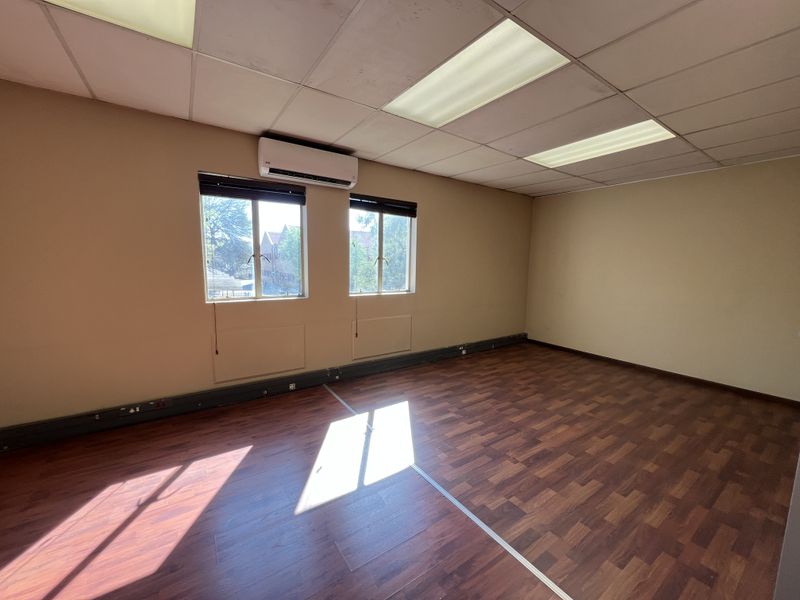 A vacant office space featuring laminate wood flooring, suspended ceiling with recessed lighting, and a wall-mounted split-system air conditioning unit.