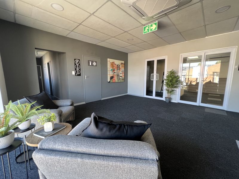 A modern office reception or waiting area featuring carpeted flooring, a suspended ceiling with integrated lighting, and glass-paneled doors leading to adjacent workspaces.