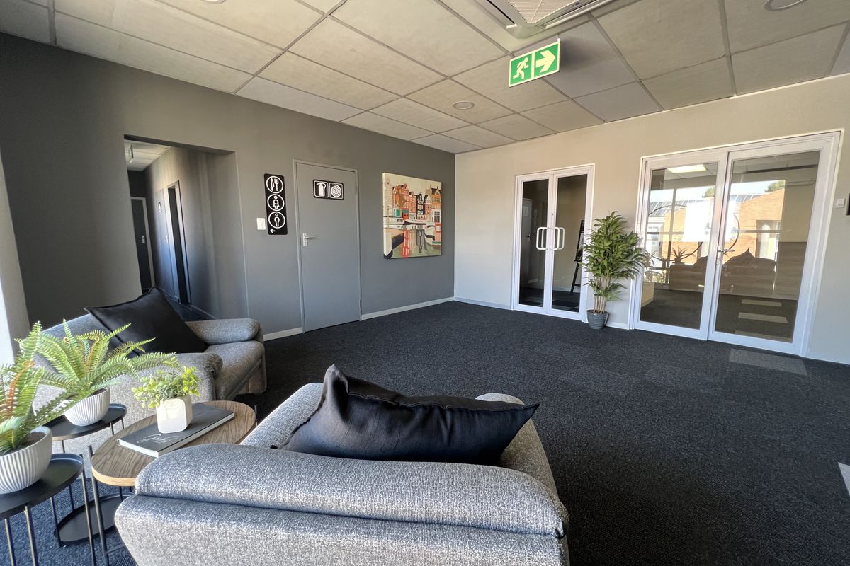 A modern office reception or waiting area featuring carpeted flooring, a suspended ceiling with integrated lighting, and glass-paneled doors leading to adjacent workspaces.