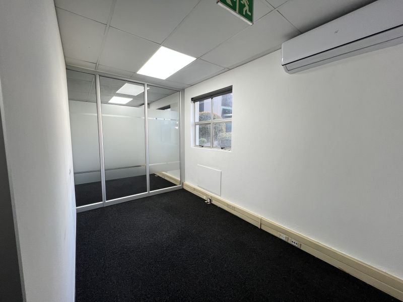 A clean, empty office space featuring a glass partition, suspended ceiling, air conditioning unit, and dark carpet flooring.