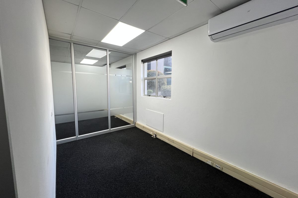 A clean, empty office space featuring a glass partition, suspended ceiling, air conditioning unit, and dark carpet flooring.