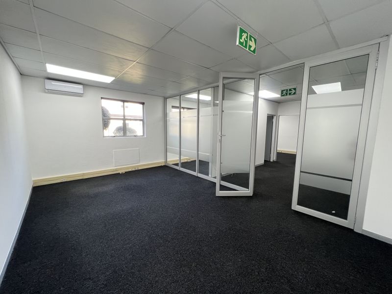 A clean, empty office space featuring glass partitions, suspended ceilings with recessed lighting, air conditioning, and dark carpeting.