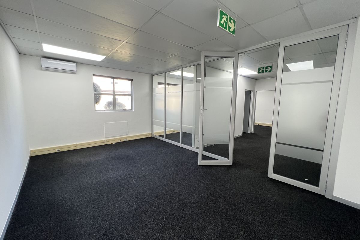 A clean, empty office space featuring glass partitions, suspended ceilings with recessed lighting, air conditioning, and dark carpeting.