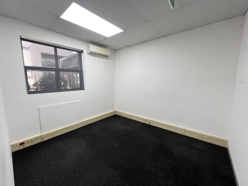 A vacant, functional office space featuring carpeted flooring, a suspended ceiling with recessed lighting, and a wall-mounted air conditioning unit.
