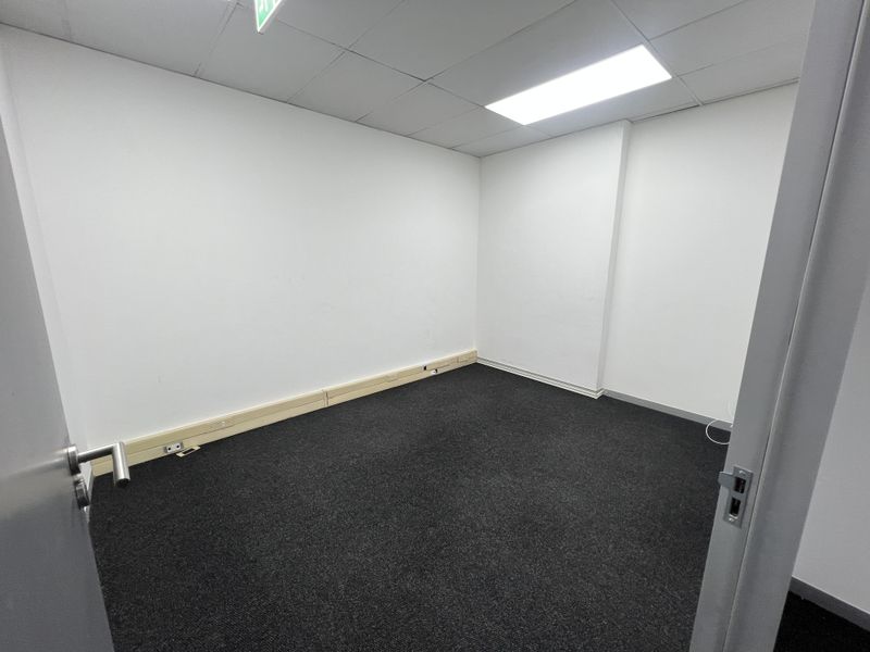 A clean, empty office space featuring dark carpet flooring, a suspended ceiling with recessed lighting, and perimeter trunking for electrical connectivity.