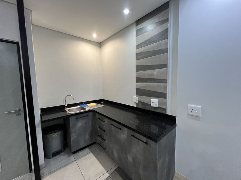 A compact office kitchenette featuring modern grey cabinetry, a black stone countertop, stainless steel sink, and decorative tiled splashback.