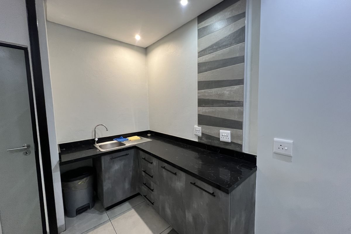 A compact office kitchenette featuring modern grey cabinetry, a black stone countertop, stainless steel sink, and decorative tiled splashback.