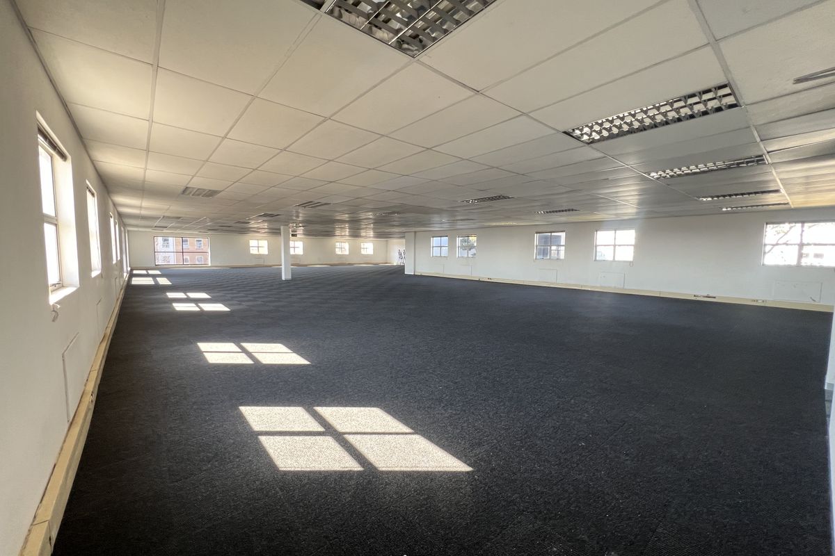 A large, open-plan office space featuring a suspended ceiling with recessed lighting, dark carpeting, and multiple windows providing natural light.