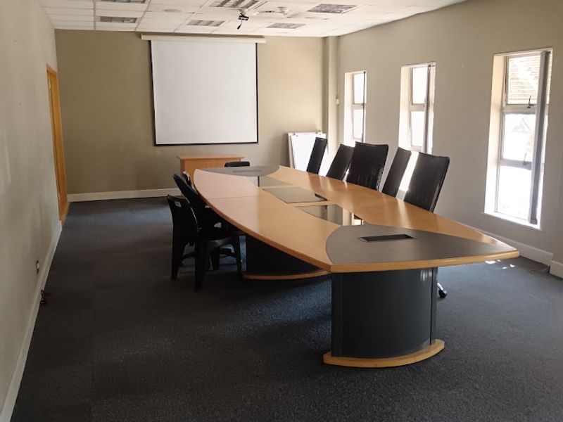 A functional office boardroom featuring a large conference table, suspended ceiling with recessed lighting, and a wall-mounted projector screen.