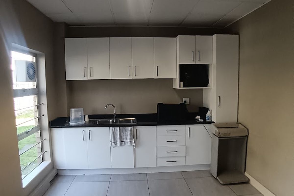 A functional office kitchenette featuring white built-in cabinetry, a stainless steel sink, tiled flooring, and a drop ceiling with integrated lighting.