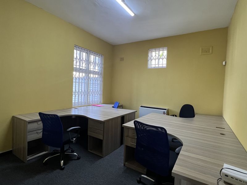 A functional office space featuring yellow walls, security bars on windows, carpeted flooring, and an integrated air conditioning unit.