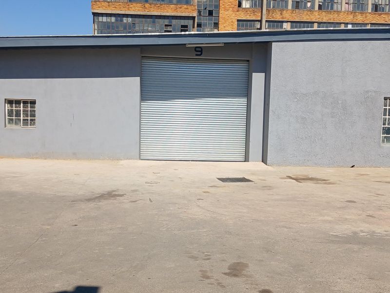 Industrial unit exterior featuring a large roller shutter door, security-barred windows, and a paved concrete apron area.